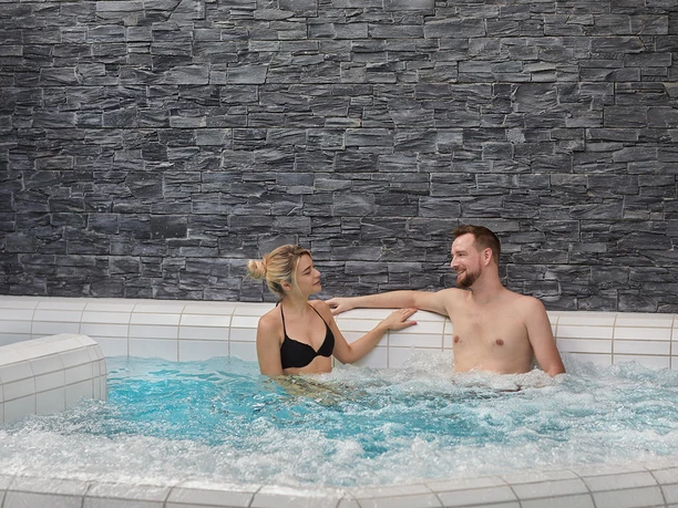 Kraeutervitalbad Sebnitz Ein Mann und eine Frau entspannen sich lächelnd in einem sprudelnden Whirlpool vor einer grauen Steinwand.