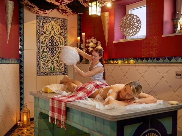 Een vrouw krijgt een schuimmassage op een marmeren tafel in een spa in oosterse stijl met rode muren en kaarslicht.