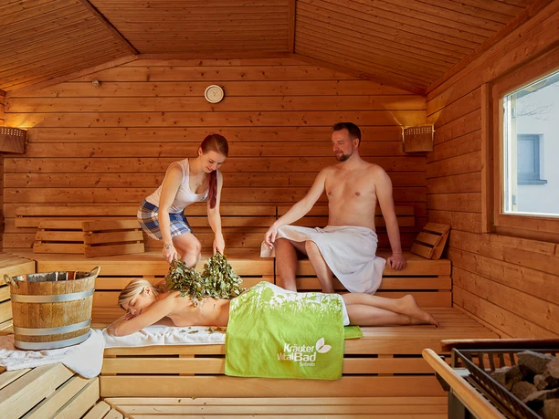 Wenik Ritual in der Sauna Drei Personen in einer Holzsauna; eine Frau liegt auf einer Bank mit einem grünen Handtuch, während eine andere Frau sie mit Zweigen massiert. Ein Mann sitzt daneben.