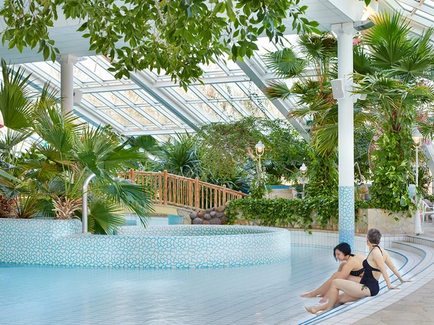 Mariba Erlebnisbad Zwei Personen in Badebekleidung sitzen am Rand eines Innenpools, umgeben von tropischen Pflanzen und einem Glasdach, das natürliches Licht hereinlässt.