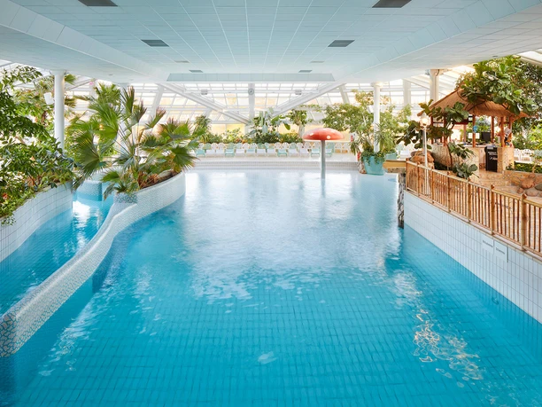 Groot, helder binnenzwembad met turquoise gekleurd water, omringd door tropische planten en een rode paddenstoelvormige waterpartij.