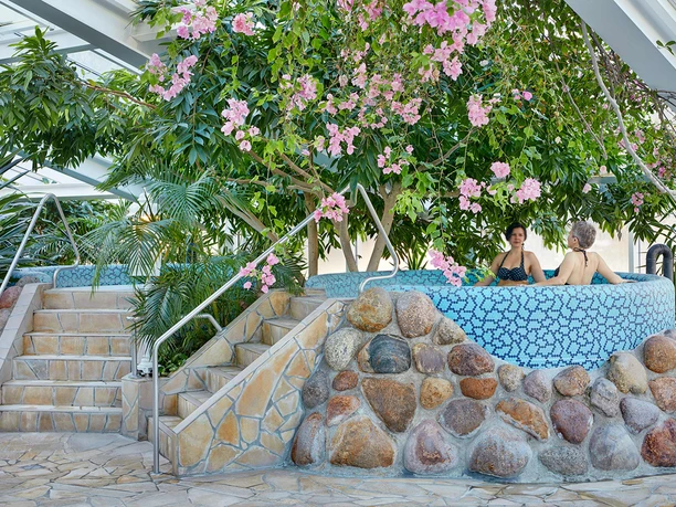 Mariba Whirlpool Zwei Personen entspannen in einem blauen Mosaik-Whirlpool, umgeben von üppigen grünen Pflanzen und rosa Blüten, mit einer ruhigen Atmosphäre.
