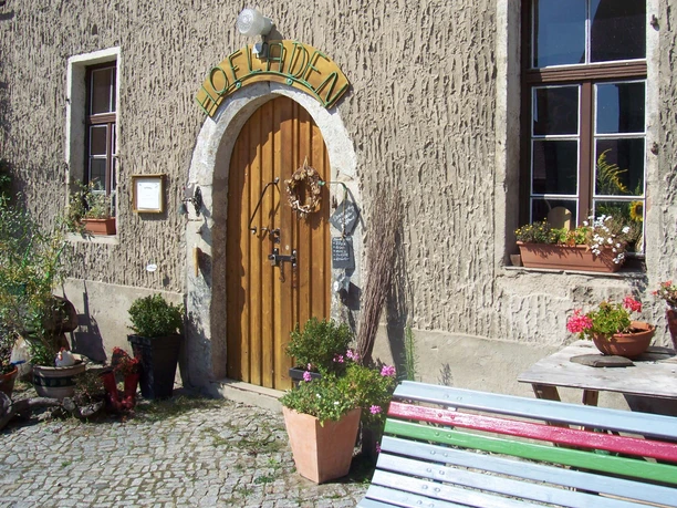 Entrance of a farm store with wooden door, surrounded by plants in pots; next to it a colorful bench and table with flower pots, sunny atmosphere.