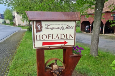A sign with the words "Hofladen" and a red arrow points the way, surrounded by green countryside and a tree.