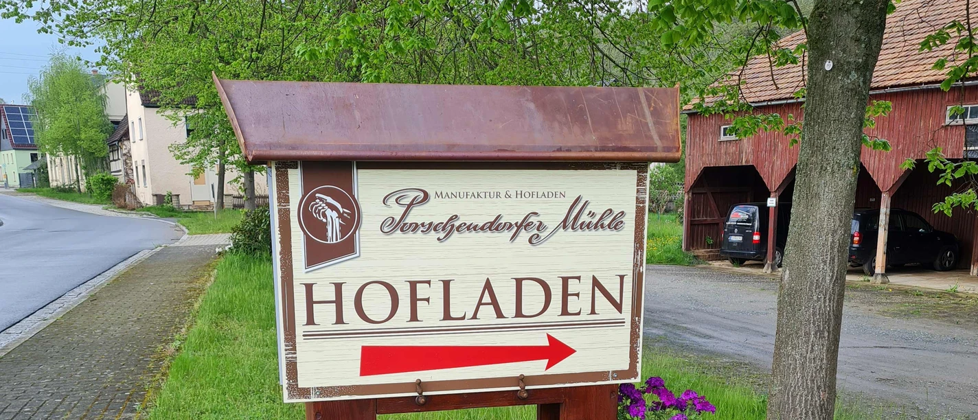 A sign with the words "Hofladen" and a red arrow points the way, surrounded by green countryside and a tree.