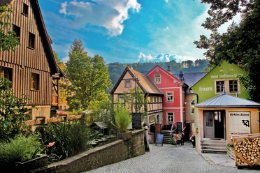 Bunte Fachwerkhäuser in einer malerischen Gasse, umgeben von grünen Bäumen und einem blauen Himmel; im Vordergrund eine Bio-Bäckerei.