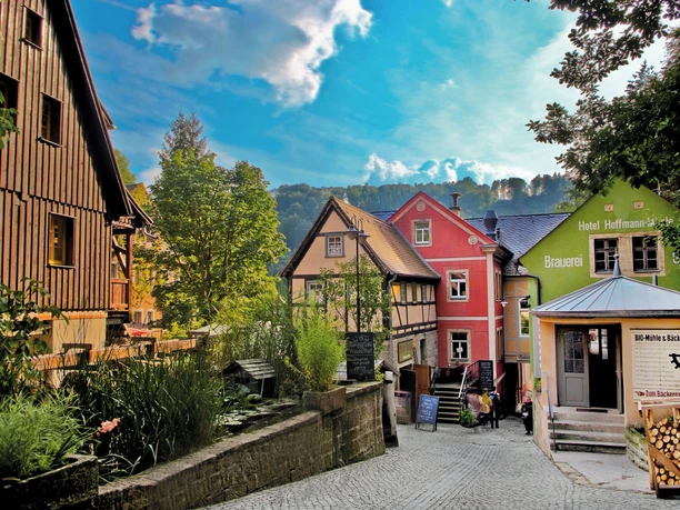 Mühlenhofensemble Schmilka Bunte Fachwerkhäuser in einer malerischen Gasse, umgeben von grünen Bäumen und einem blauen Himmel; im Vordergrund eine Bio-Bäckerei.