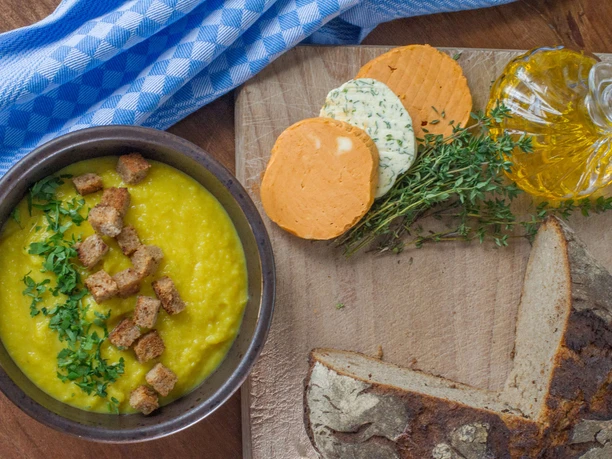 Mühlenhof Ausschank Schmilka Gelbe Suppe mit Croutons und Petersilie in einer Schüssel, daneben Brot, Kräuterbutter und Olivenöl auf einem Holzbrett, mit blau-weißem Tuch.