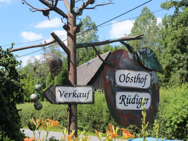 Metal sign in the shape of an apple with the inscription "Obsthof Rüdiger" and "Verkauf", surrounded by green vegetation and flowering lilies.