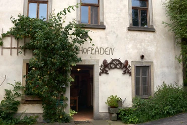 Oud gebouw met het opschrift "Hofladen", omgeven door klimplanten; links een open raam, rechts een deur met planten ervoor.
