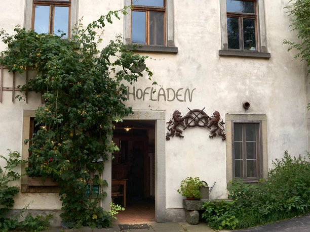 Old building with the inscription "Hofladen" (farm store), surrounded by climbing plants; on the left an open window, on the right a door with plants in front of it.