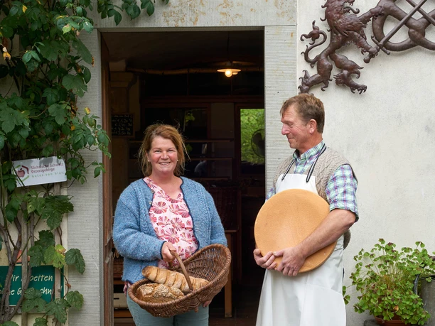 Een man en een vrouw staan glimlachend voor een gebouw; de vrouw houdt een mand brood vast, de man een groot wiel kaas.
