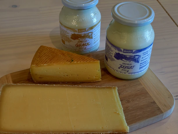 Angebot Bauernhof Steinert Ein Holzbrett mit einem Stück gelbem Käse und zwei Gläsern Demeter-Quark und -Joghurt auf einem hellen Holztisch.