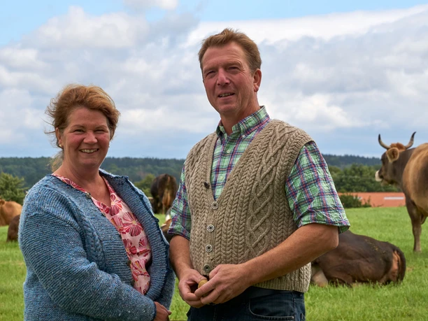 Bauernhof Steinert Ein Mann und eine Frau stehen lächelnd auf einer grünen Wiese mit Kühen im Hintergrund, unter einem bewölkten Himmel.