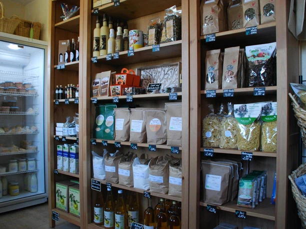 Hausgemachte Fruchtaufstriche Regale mit verschiedenen Lebensmitteln, darunter Nudeln, Mehl und Flaschen, neben einem Kühlschrank mit Milchprodukten in einem kleinen Laden.