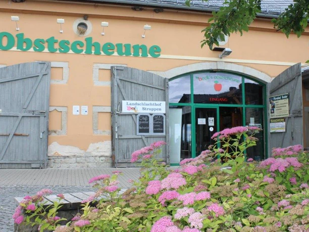 Obstscheune-Krietzschwitz-3.jpg Eingang der Obstschenke mit grüner Schrift auf orangefarbener Fassade, umgeben von blühenden rosa Blumen und offenen Holztüren.