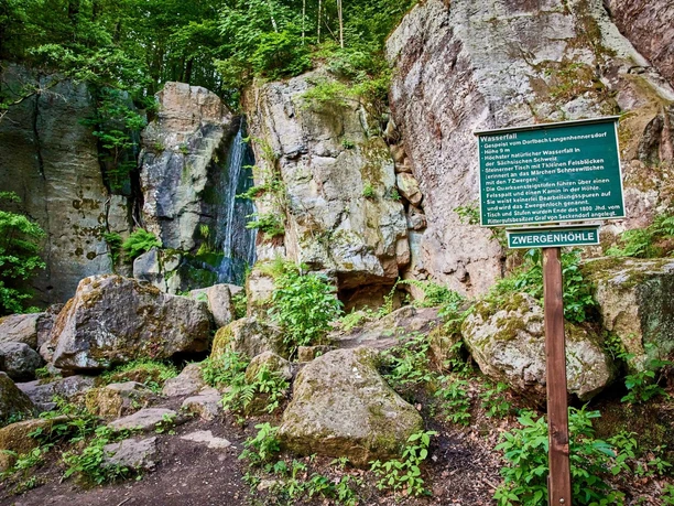 Rocky landscape with a small waterfall, surrounded by green vegetation; a sign with information about the "Dwarf Cave" stands in the foreground.