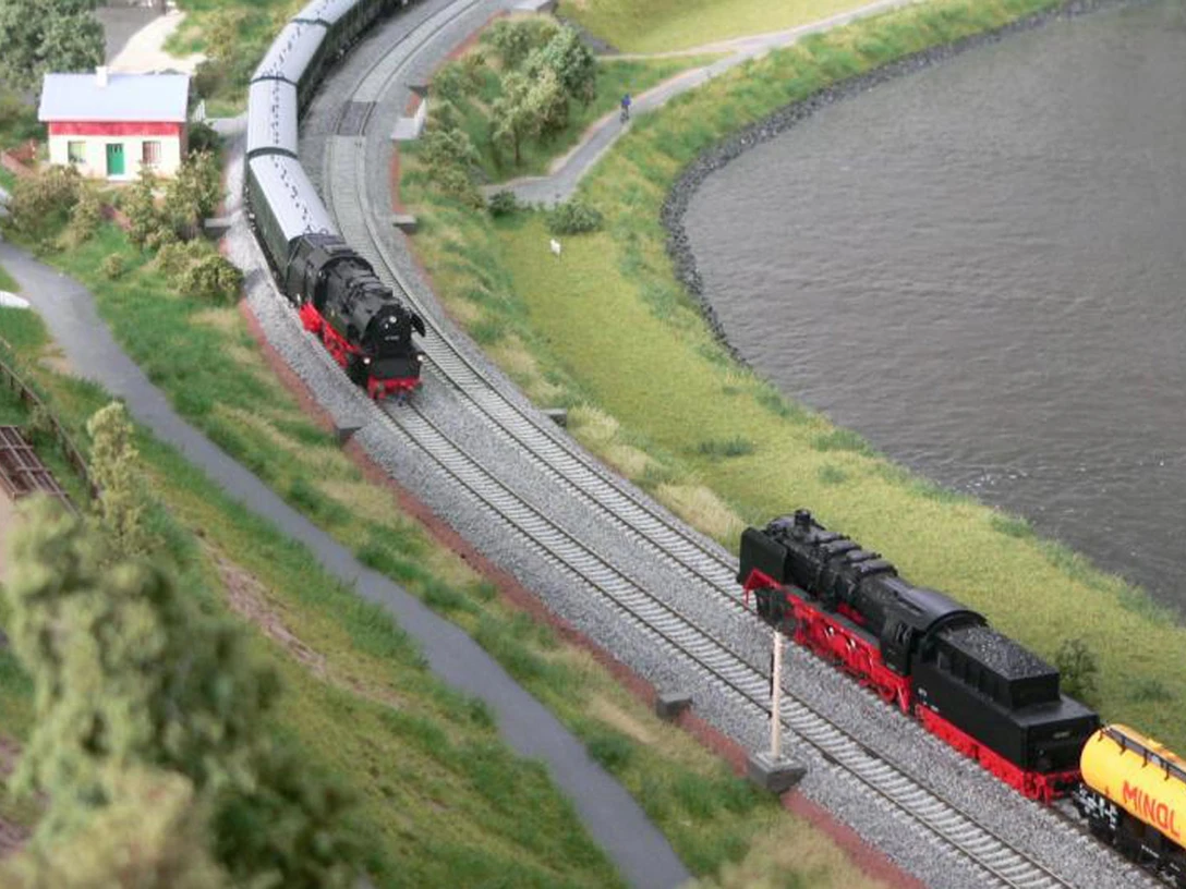 Miniatur Elbtal Modellbahnservice Königstein Modelleisenbahn-Szene mit zwei schwarzen Dampflokomotiven auf parallelen Gleisen, umgeben von grüner Landschaft und einem Gewässer.
