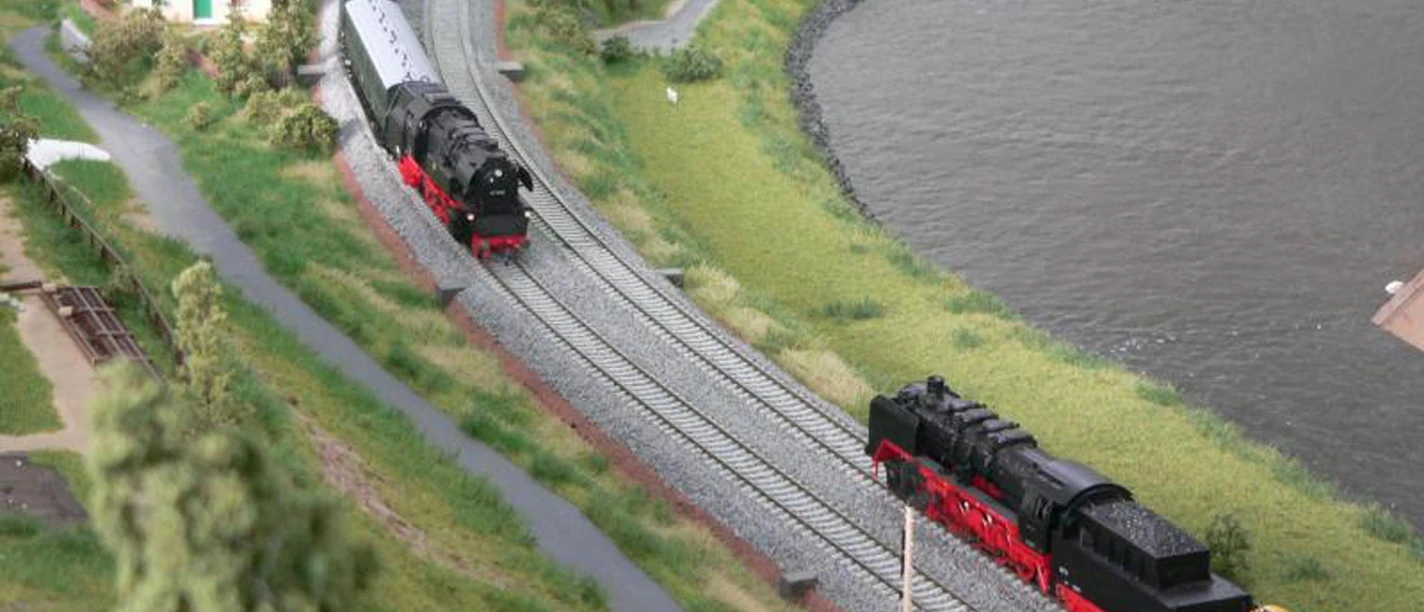 Miniatur Elbtal Modellbahnservice Königstein Modelleisenbahn-Szene mit zwei schwarzen Dampflokomotiven auf parallelen Gleisen, umgeben von grüner Landschaft und einem Gewässer.