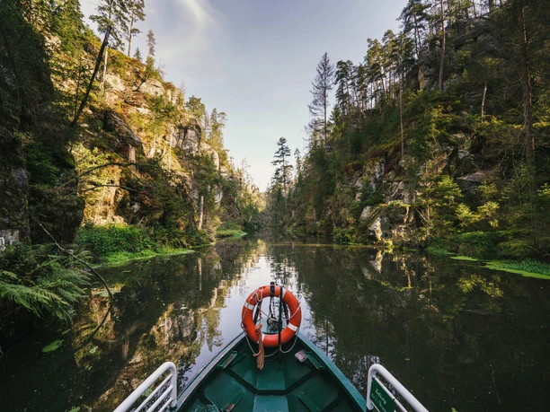 A green boat with a lifebuoy sails through a narrow gorge lined with trees and rocks that are reflected in the calm water.