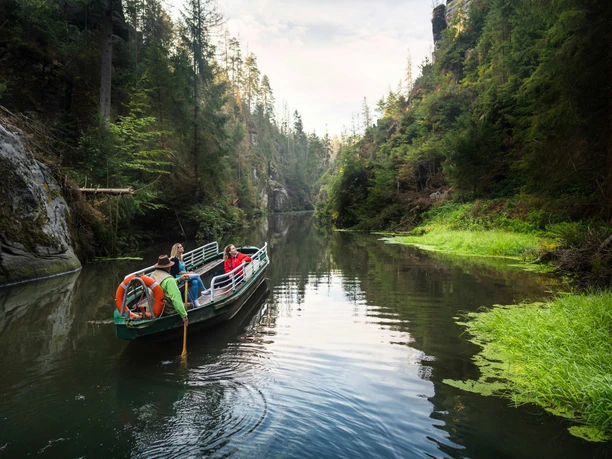 A boat with three people sails through a quiet, wooded gorge with green water and steep rock faces.