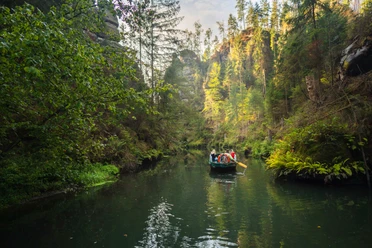 A group of people are traveling in a boat on a calm river, surrounded by dense, green forest and steep rock faces.
