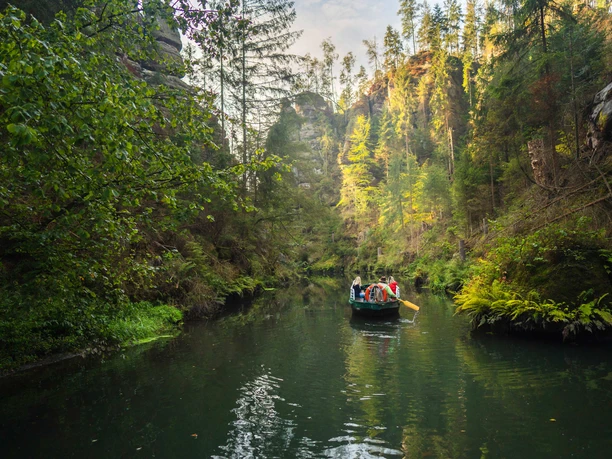 A group of people are traveling in a boat on a calm river, surrounded by dense, green forest and steep rock faces.