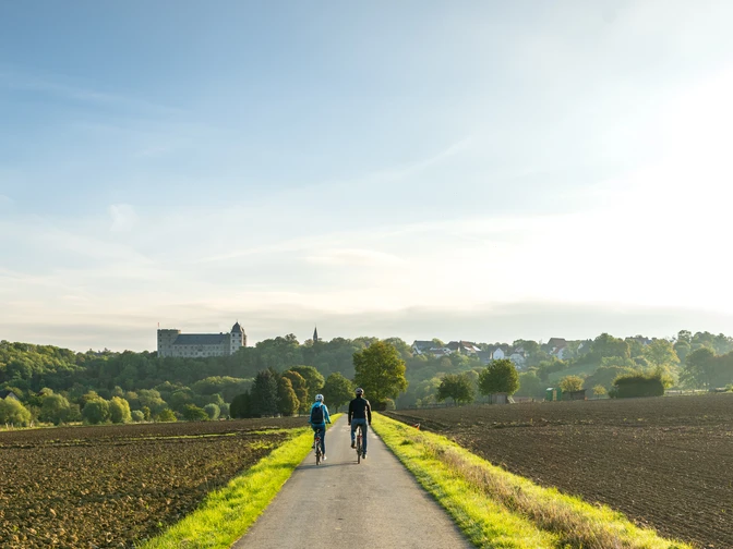 Blick auf drei Radfahrer auf einem ländlichen Weg, umgeben von Feldern und Bäumen, mit Burg im Hintergrund.