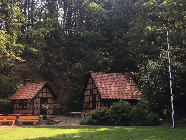 Zwei traditionelle Fachwerkhäuser mit roten Ziegeldächern stehen umgeben von Bäumen im Wald.