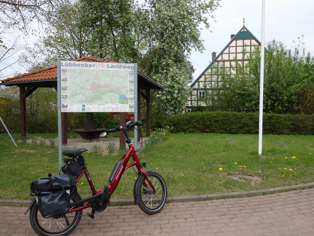 Fahrrad steht vor Infotafel des Lübbecker Landwegs, umgeben von Bäumen und einem Fachwerkhaus.