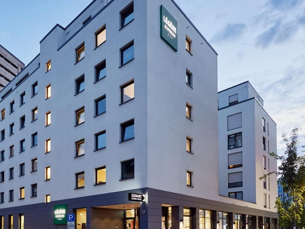 Ein modernes Gebäude mit klaren Linien und Fenstern. Die Fassade ist hell und freundlich, umgeben von Pflanzen.