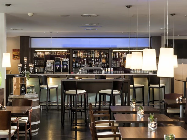 Ein modernes Restaurant mit einer eleganten Bar im Hintergrund. Helle Beleuchtung und stilvolle Möbel schaffen eine einladende Atmosphäre.