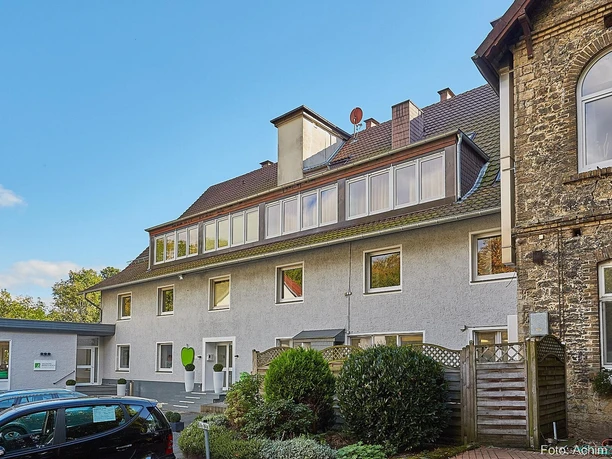 Ein modernes Gebäude mit mehreren Fenstern und einem begrünten Bereich davor. Nebenan steht ein älteres, rustikales Gebäude aus Stein.