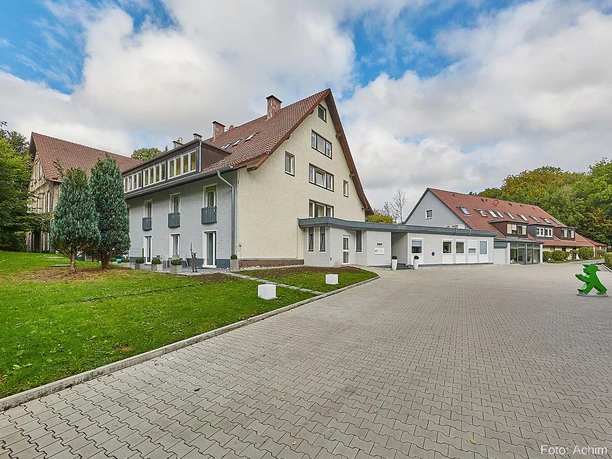 Ein modernes Gebäude in einer ruhigen Umgebung mit Grünflächen und Bäumen. Der gepflasterte Weg führt zum Eingang des Hauses.