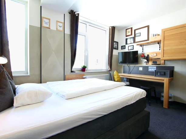 Ein modernes Hotelzimmer mit einem Doppelbett und einem Schreibtisch. Die Wände sind mit Bildern dekoriert und es gibt viel Tageslicht.
