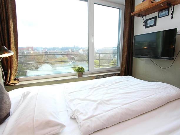 Ein gemütliches Schlafzimmer mit einem großen Fenster und Blick auf eine Stadtlandschaft. Die Einrichtung ist modern mit einem Fernseher und einem dekorativen Regal.