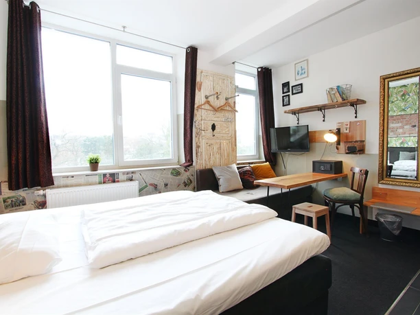 Ein modernes Zimmer mit einem großen Fenster und hellen Vorhängen. Das Zimmer verfügt über ein Bett, einen Schreibtisch und eine gemütliche Sitzecke.