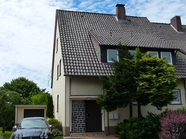 Ein modernes Haus mit einem begrünten Garten und einer Zufahrt. Ein schwarzes Auto steht vor der Haustür auf einem gepflasterten Weg.