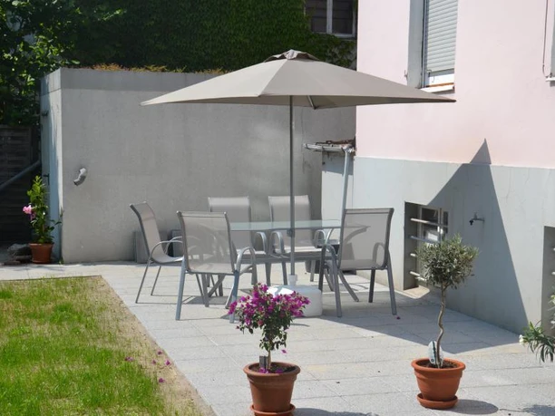Ein gemütlicher Gartentisch mit Stühlen und einem Sonnenschirm steht auf einer Sonnenterrasse. Im Hintergrund gibt es blühende Pflanzen und gepflegte Rasenfläche.