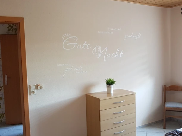 Schlafzimmer 1.jpg Schlafzimmer mit hellem Holzschrank, dekorativer Schrift an der Wand und gefliesten Bodenfliesen.