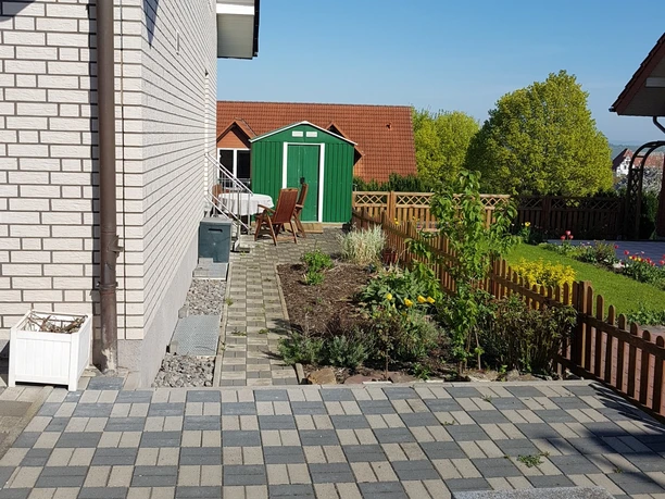 Terasse Sitzecke Fewo.jpg Sitzbereich auf gepflasterter Terrasse neben einem kleinen Garten mit Holzzaun, im Hintergrund grüne Natur.