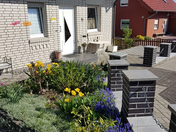 Eingangsbereich eines Hauses mit Blumenbeet, Sitzgelegenheiten und gepflastertem Weg im Vordergrund.