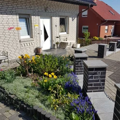 Aussenansicht Eingangsbereich Eingangsbereich eines Hauses mit Blumenbeet, Sitzgelegenheiten und gepflastertem Weg im Vordergrund.