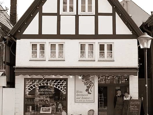 Fachwerkhaus mit großem Giebeldach, zwei Menschen stehen neben Schaufenster mit Weinaufdruck, Gehweg davor.