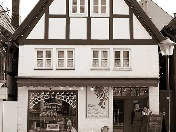 Fachwerkhaus mit großem Giebeldach, zwei Menschen stehen neben Schaufenster mit Weinaufdruck, Gehweg davor.