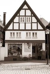 hausanpl.jpg Fachwerkhaus mit großem Giebeldach, zwei Menschen stehen neben Schaufenster mit Weinaufdruck, Gehweg davor.