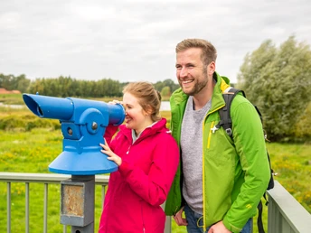 Paar in bunter Outdoor-Kleidung blickt durch blaues Fernrohr auf Landschaft von einer Aussichtsplattform.