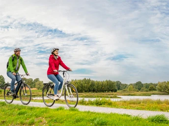 <p>Zwei Menschen radeln auf einem idyllischen Weg entlang eines Sees bei bewölktem Himmel.</p>