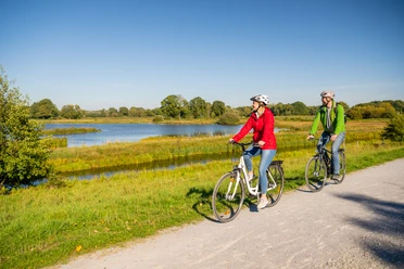 Delbrueck-Steinhorster Becken-Teutoburger-Wald-Tourismus-D-Ketz-098.jpg Zwei Radfahrer auf Schotterweg entlang eines Sees bei Sonnenschein; grüne Landschaft im Hintergrund.