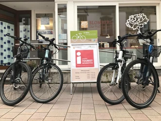 IMG_0570.jpg Vier E-Bikes stehen auf einer gepflasterten Fläche vor einem Tourist-Informationszentrum.